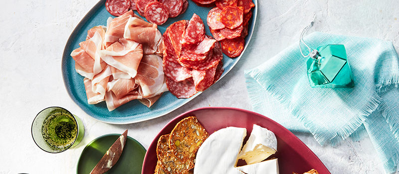 Blue plate with salami and other cured meats; pink board with cheese and crackers with side plates and glasses.