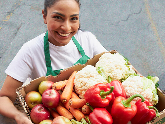 A woman working with Second Harvest is looking up to the camera with a smile on her face as she holds a large cardboard box filled with red peppers, cauliflour, carrots, and apples that have been rescued from waste.