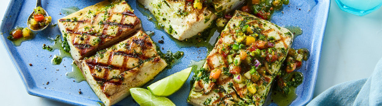 Mahi Mahi filets with a summer herb sauce and heirloom tomato salsa on a periwinkle plate with lime segments scattered on platter.