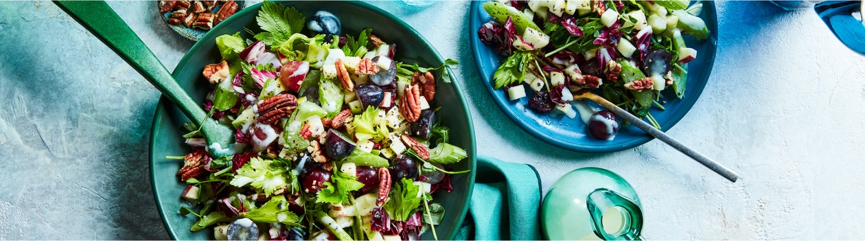 Large blue aqua-green bowl of waldorf-style salad with pecan garnish, alongside small blue side plate with single serving of salad and a side carafe of salad dressing.