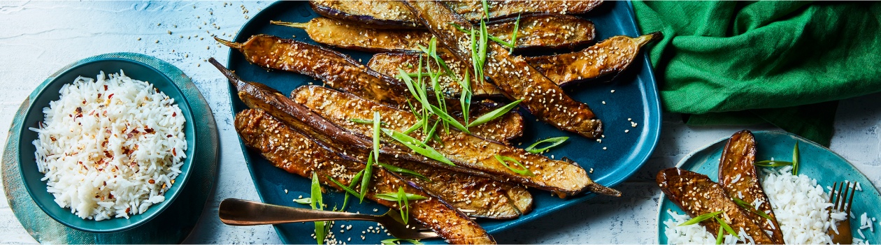 Rectangular blue serving platter with Japanese eggplant that has been roasted and garnished with fresh green onion and toasted sesame seeds with a side of white rice in a bowl