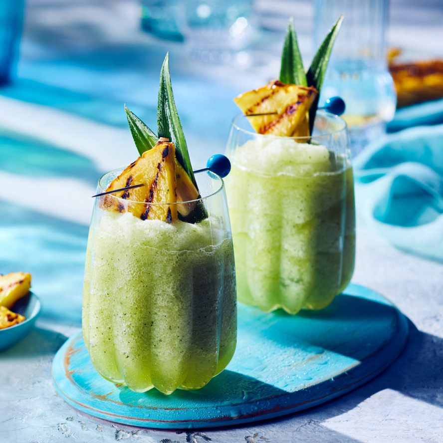 Two clear lowball glasses with green slushy Pina Vida drink, garnished with pineapple fronds