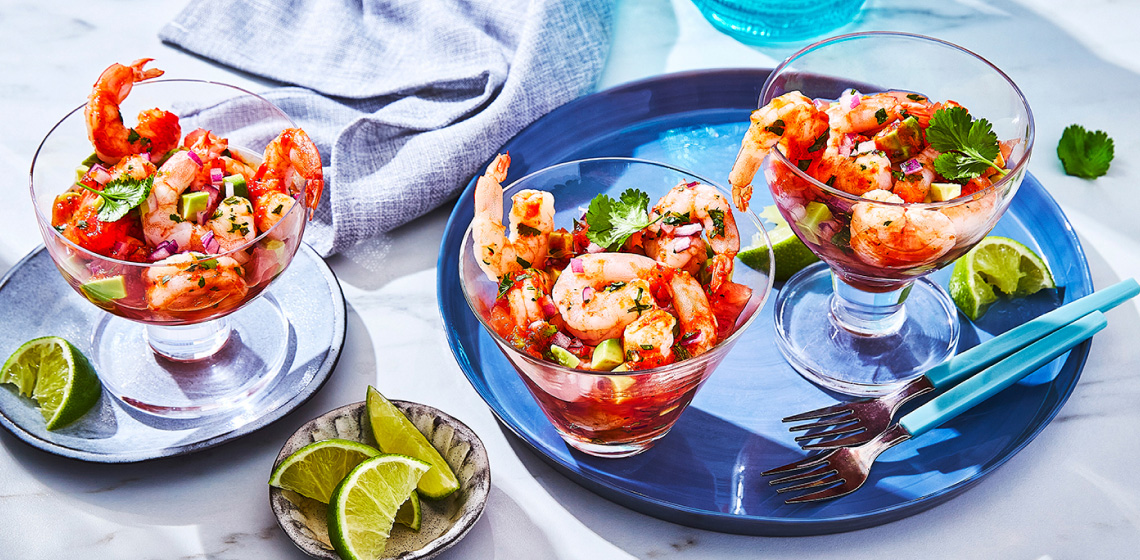 Three glass cups of Spanish-style shrimp cocktail on plates with lime wedges and cilantro for garnish.