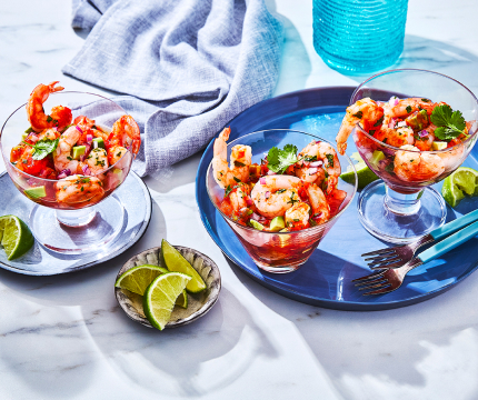 Three glass cups of Spanish-style shrimp cocktail on plates with lime wedges and cilantro for garnish.