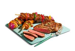 Cutting board of cooked striploin steak, partially sliced, with variety of kabobs in the background