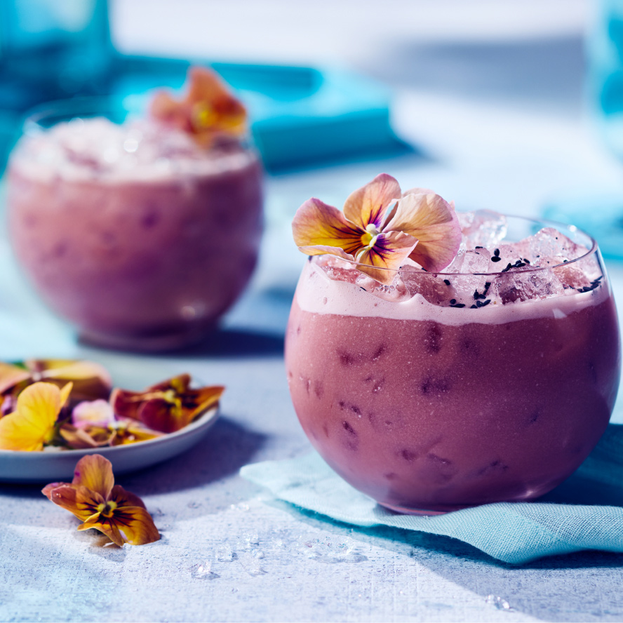 Low rounded glass of purple iced cocktail with edible floral garnish