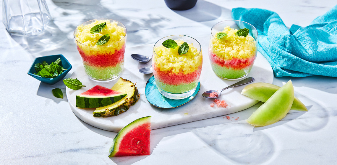 Three lowball glasses of frozen layered melon granita on a serving board with slices of melon in the foreground