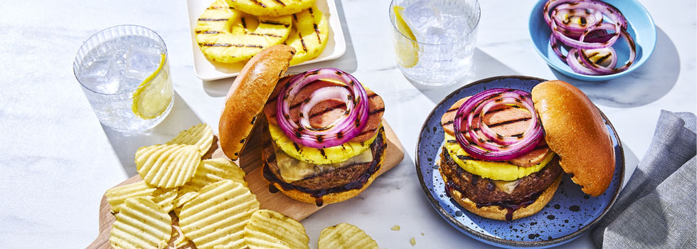 Two Hawaiian-inspired burgers with grilled pineapple rounds and a side of ruffled plain potato chips