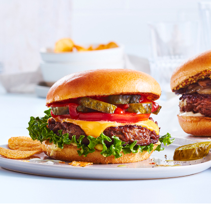 White dinner plate with a loaded beef burger, dill pickle spear and side of thick-cut fries.