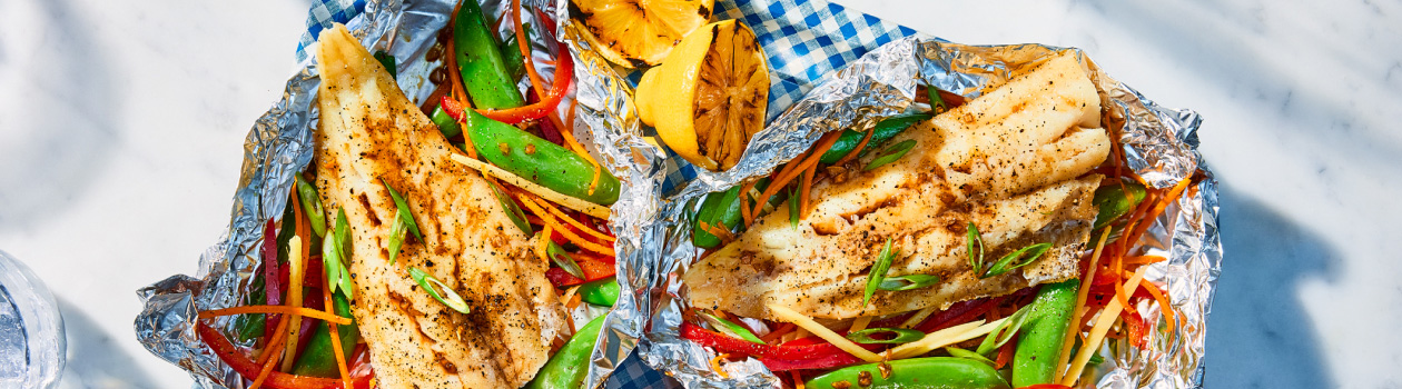 Platter with two cooked white fish fillets in foil with vegetables and grilled lemon